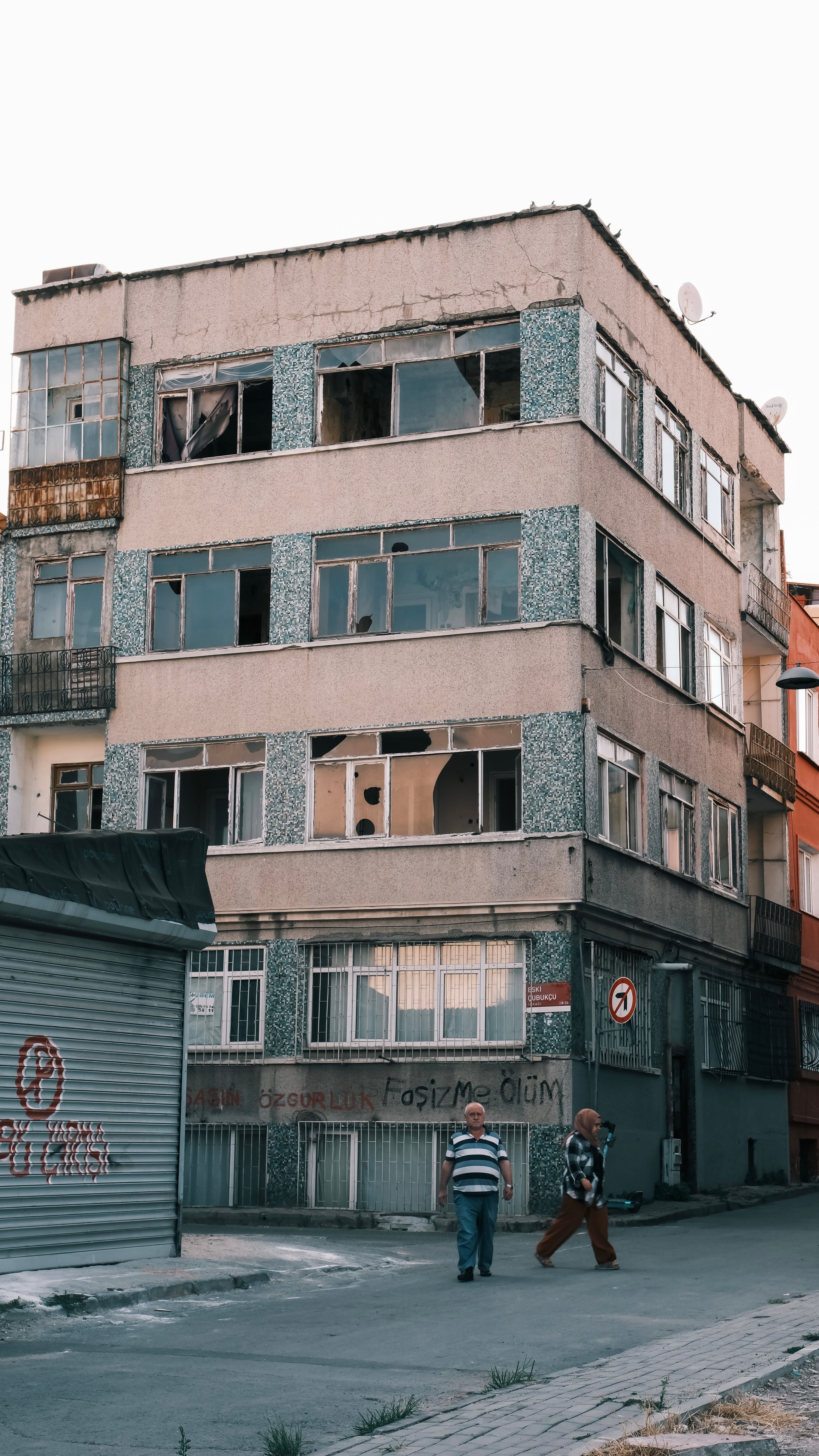 A weathered multi-story urban building with broken windows and two pedestrians on a city street.