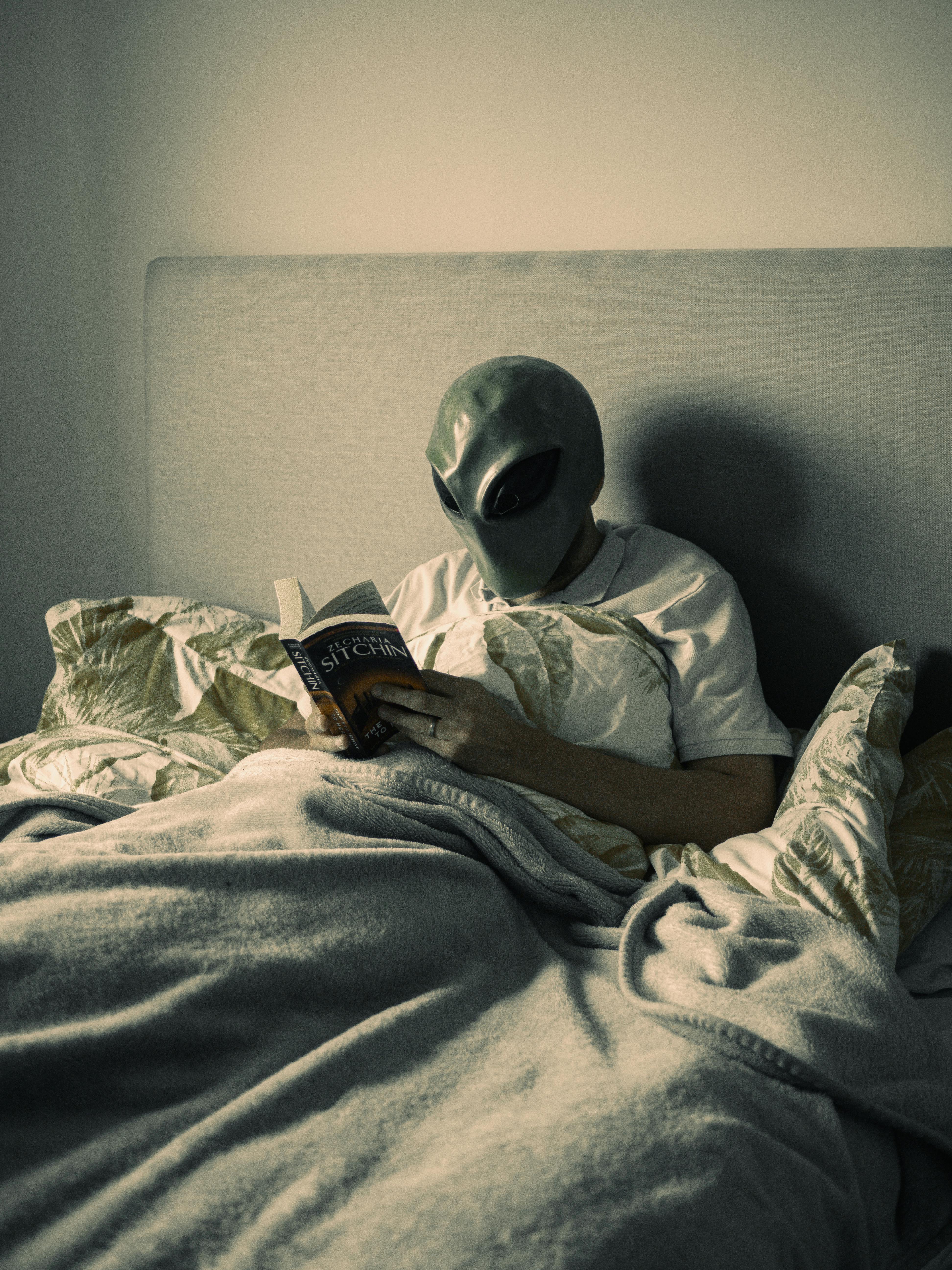 person in alien mask sitting in bed and reading book