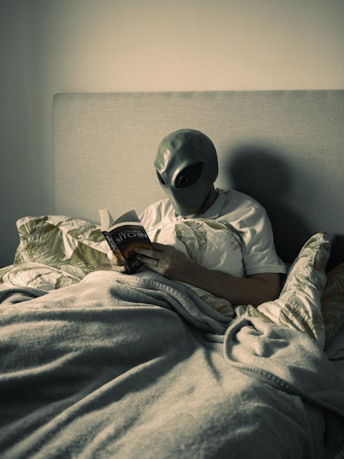 Free Person in Alien Mask Sitting in Bed and Reading Book Stock Photo