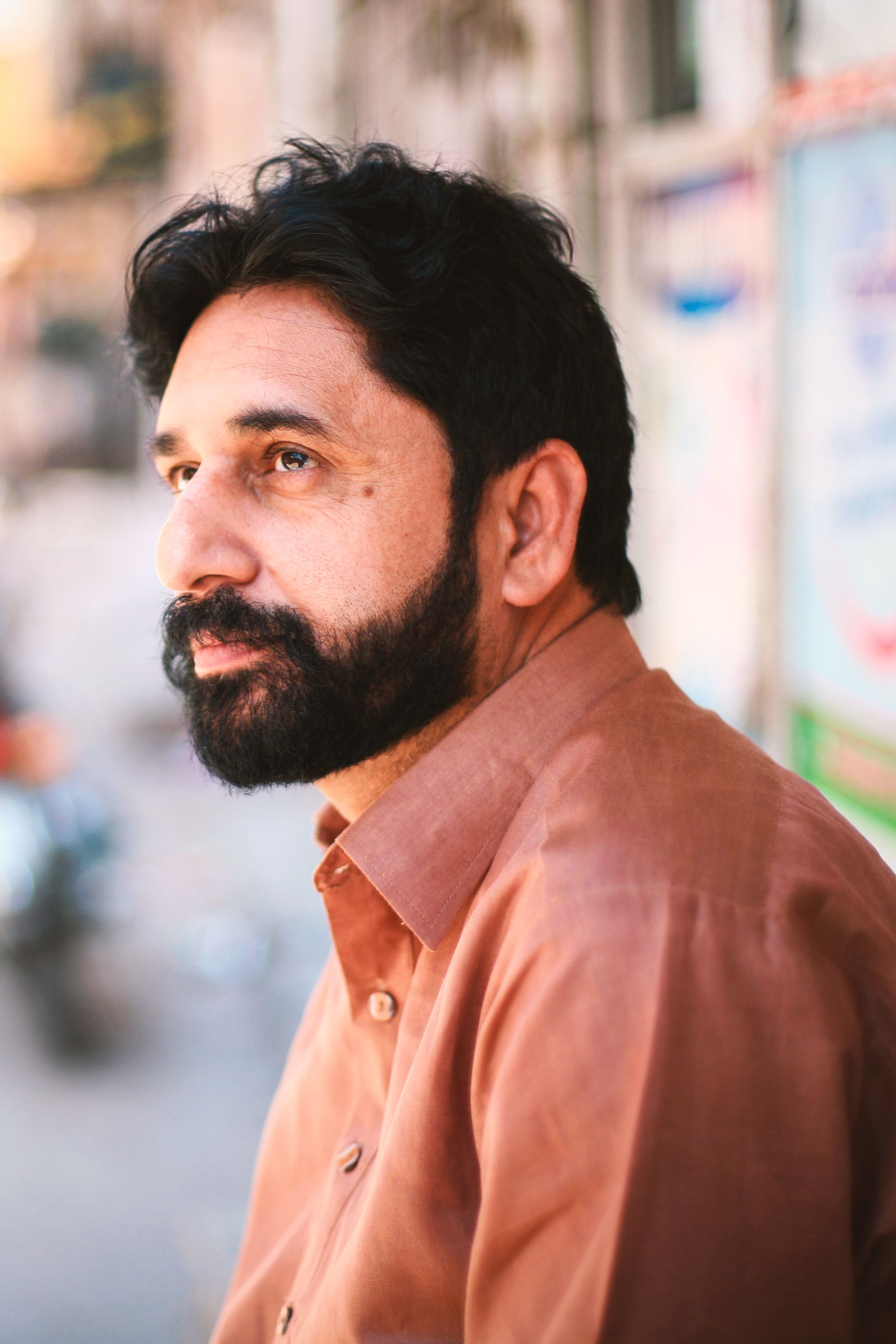 Close-up portrait of a bearded adult man with short hair in an urban outdoor setting.