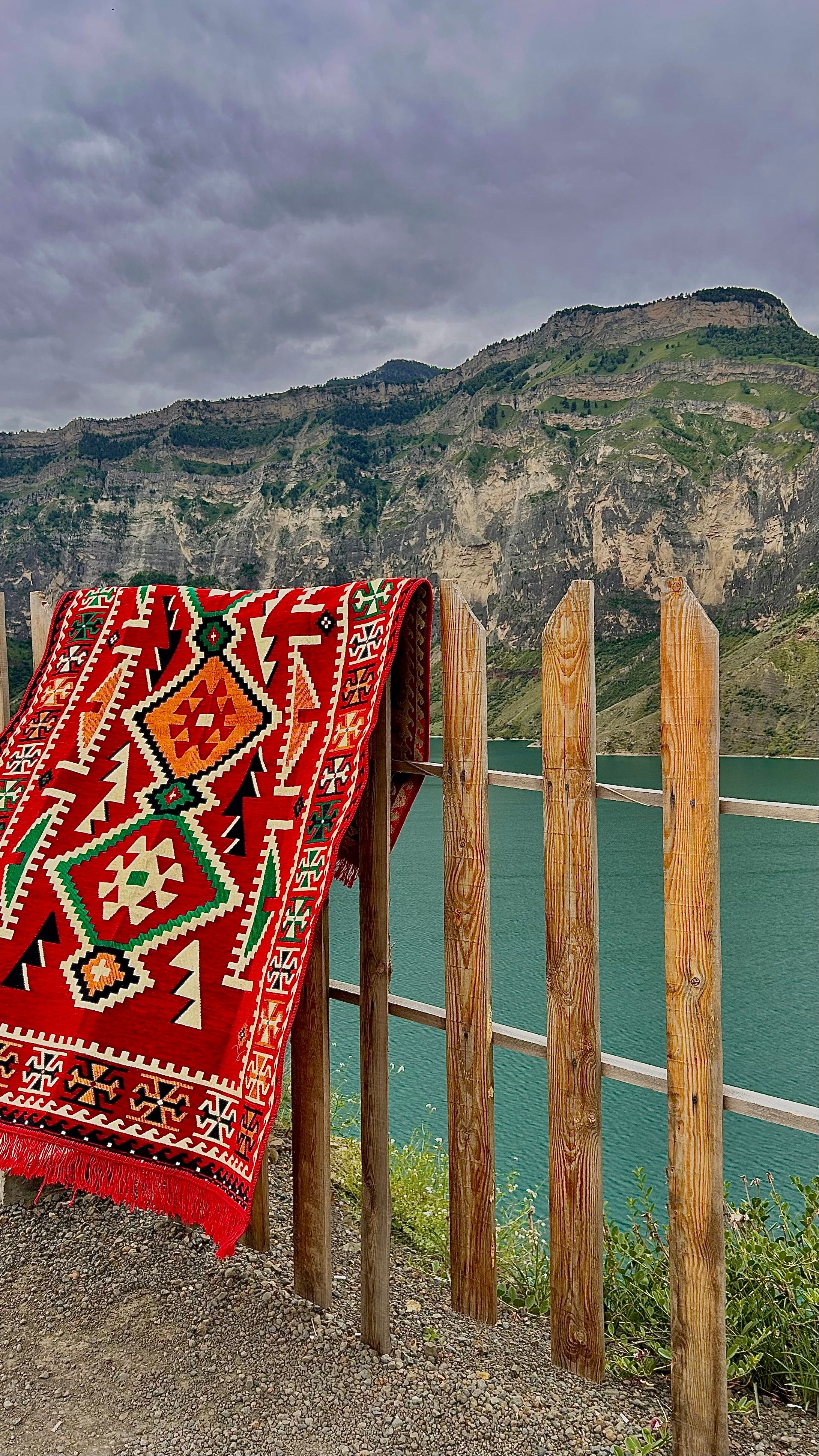 Brightly patterned rug hangs on a fence overlooking a serene lake and mountains.