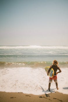 A young surfer holding his board while looking at the ocean waves, perfect day for a surf adventure.