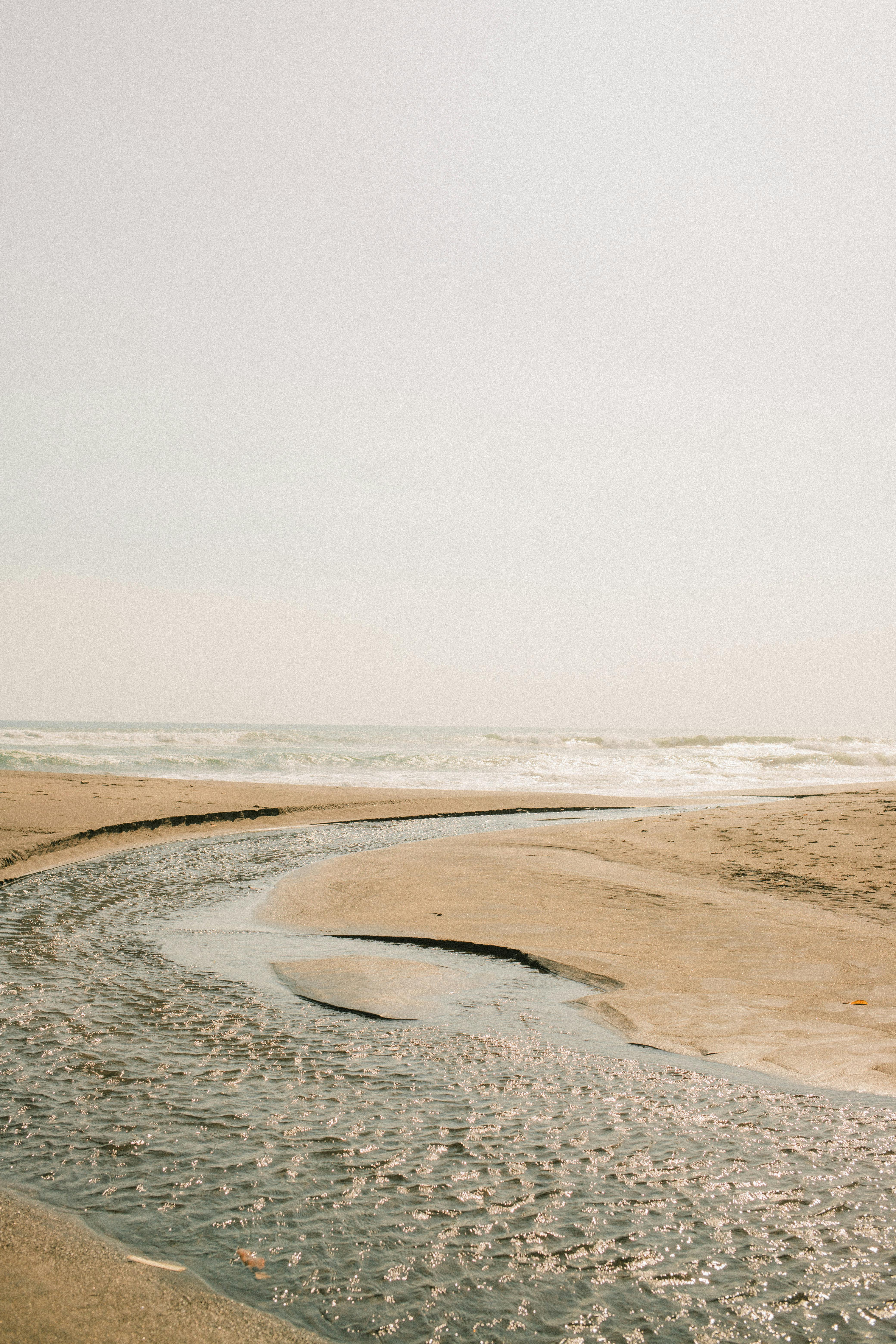 Serene beach scene with a winding stream leading to the sea, under a clear sky.