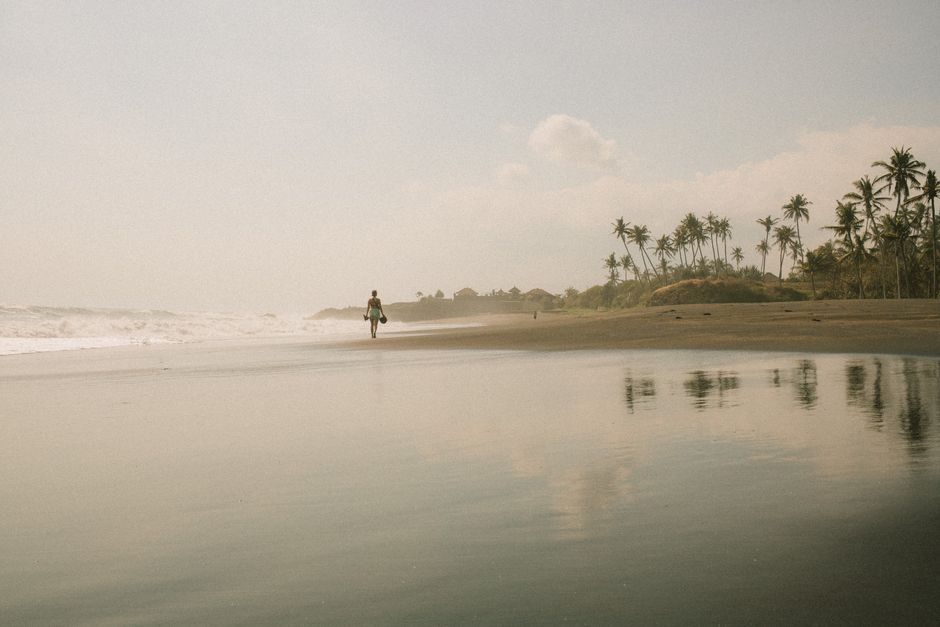 A solitary figure walks along a tranquil tropical beach at sunset, with palm trees lining the shore.