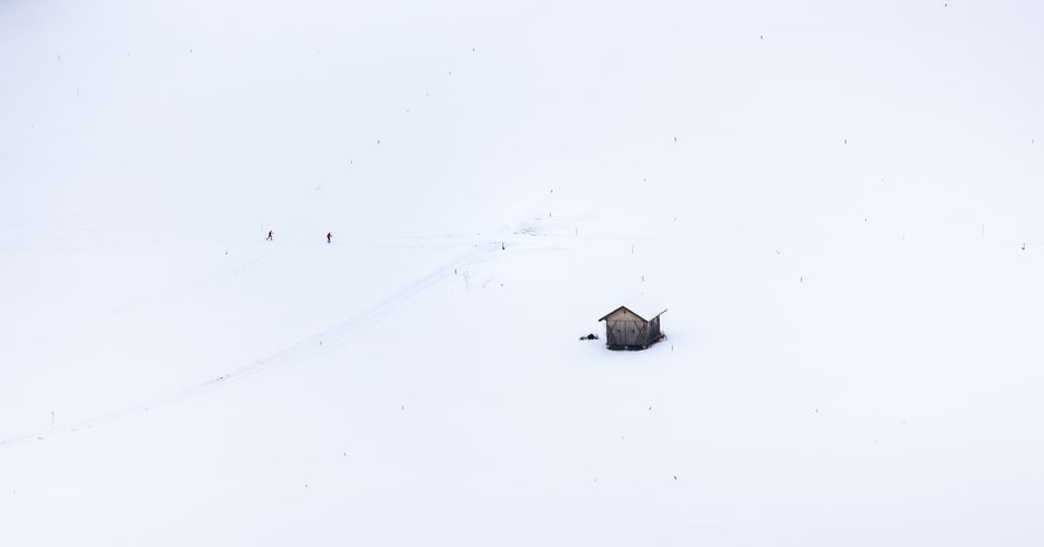 A serene snow-covered landscape featuring skiers and a solitary cabin amidst vast whiteness.
