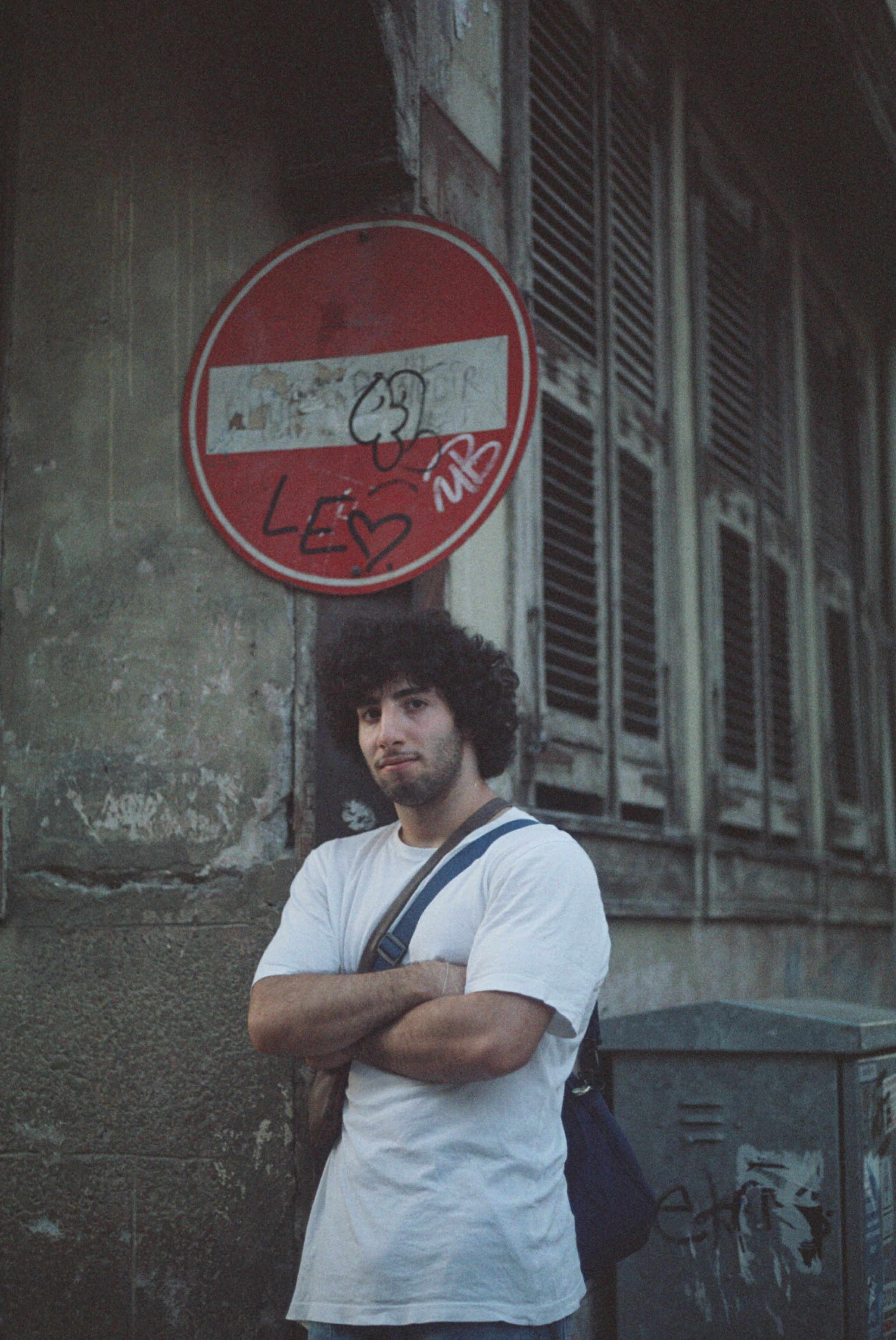A man with curly hair stands confidently by a graffitied road sign on an urban street.
