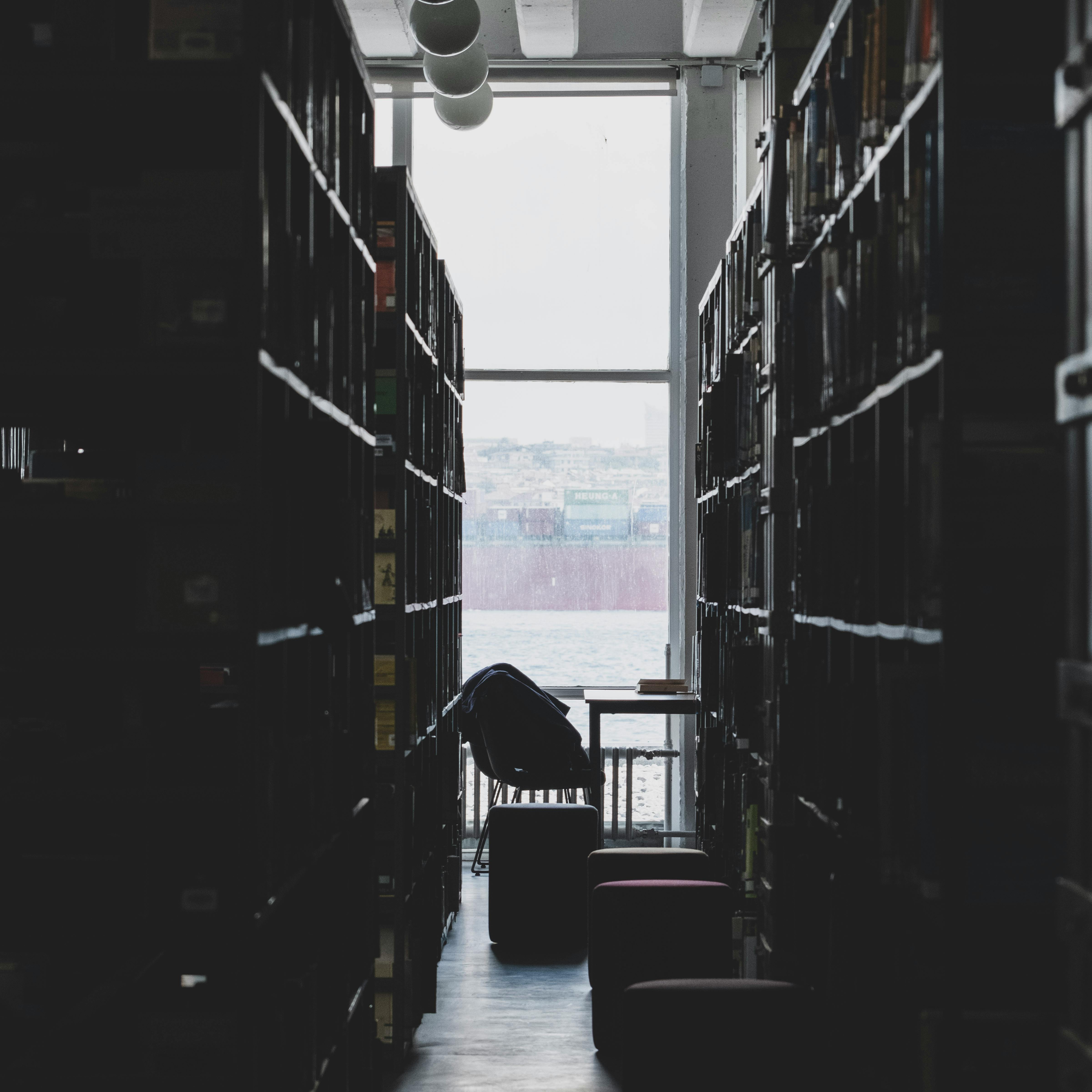 Dimly lit library aisle leading to a large window overlooking the sea. Serene and contemplative atmosphere.