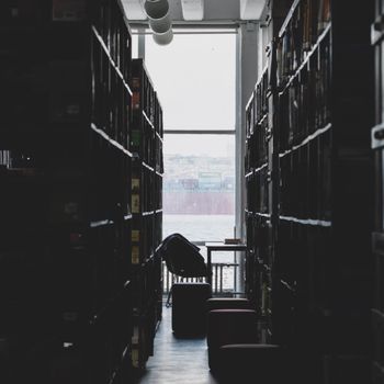 Dimly lit library aisle leading to a large window overlooking the sea. Serene and contemplative atmosphere.（イメージ）