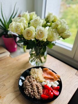 A nutritious meal with fresh ingredients and a vase of white roses by a window.