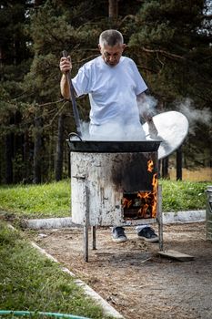 Man cooking outdoors over open flame with steam rising on a sunny day.