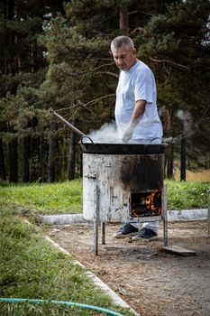 Middle-aged man cooking on a rustic outdoor stove in a forest setting, creating an authentic barbecue experience.