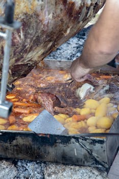 Close-up of a barbecue grill with meat and potatoes being cooked outdoors. Man's hand visible.