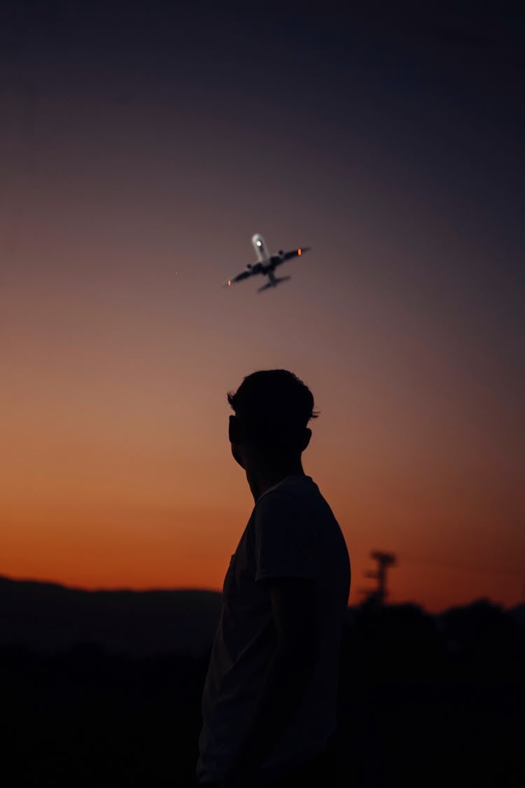 Silhouette Of A Man Standing Outside At Sunset And Looking At A Flying Airplane 