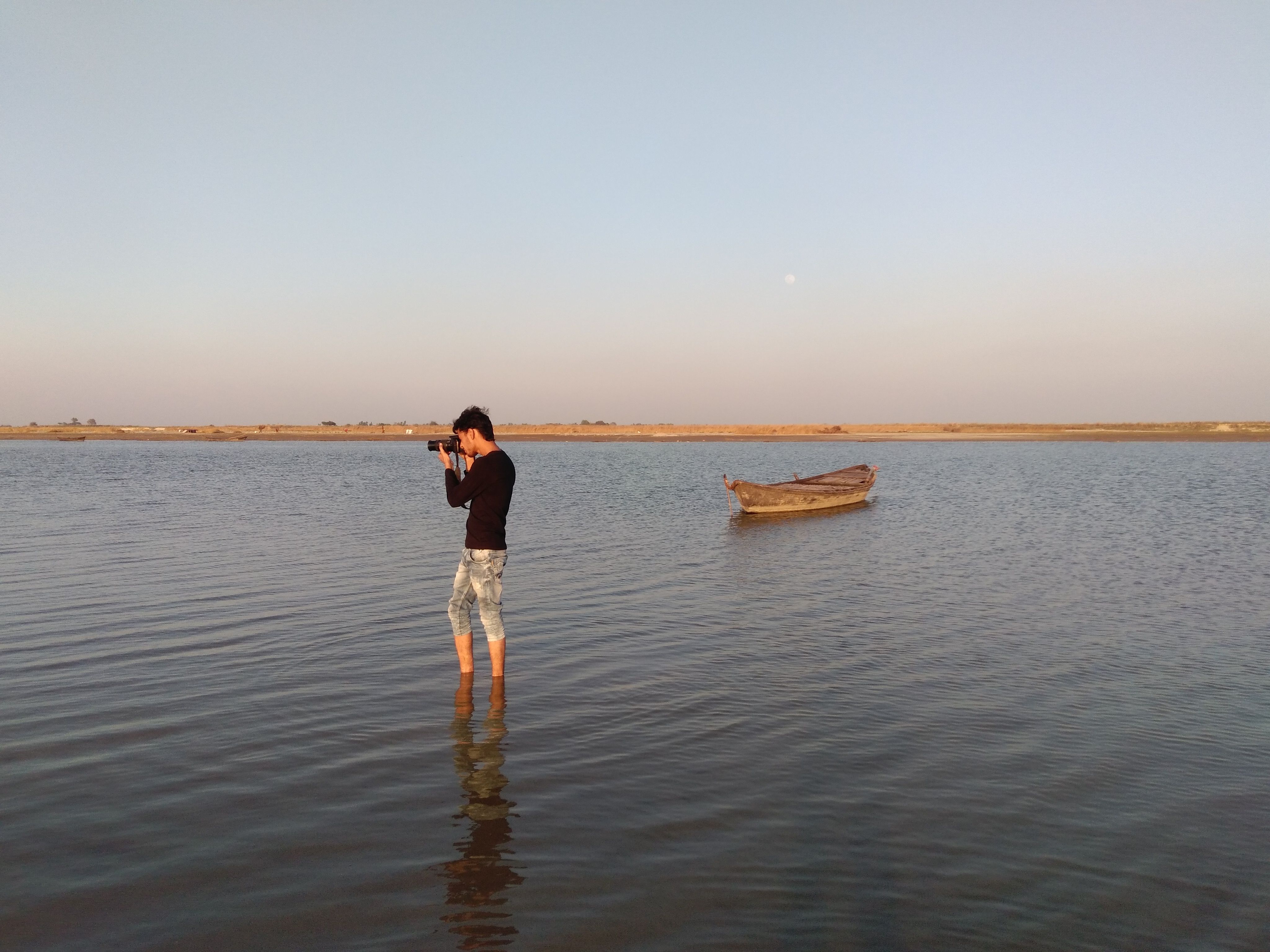 A man stands in water photographing a serene lake with a boat at Majha Chhajjapur.