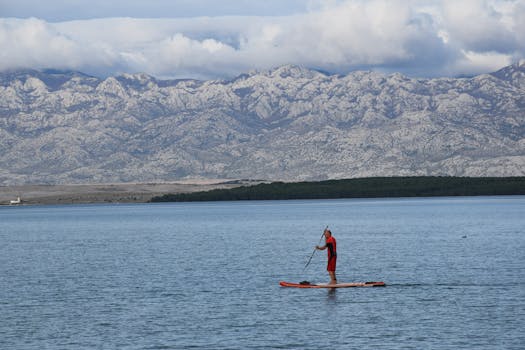 A man paddleboards on a serene lake with stunning mountain and cloud backdrop. Adventure awaits.