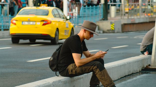A man in a hat sitting on a city street kerb using a smartphone.
