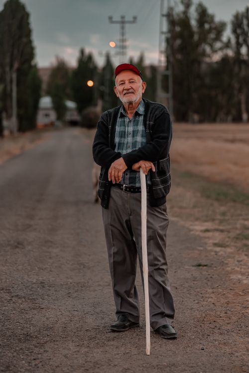 Free Senior man with a red cap and cane standing on a rural dirt road in İlkindi, Sivas, Türkiye. Stock Photo