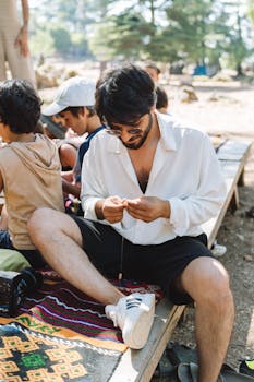 A man in a white shirt sits outdoors with friends on a sunny day, crafting while relaxing.