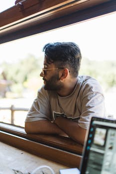Man with sunglasses and short hair gazing out of a window with arms crossed, next to a laptop.