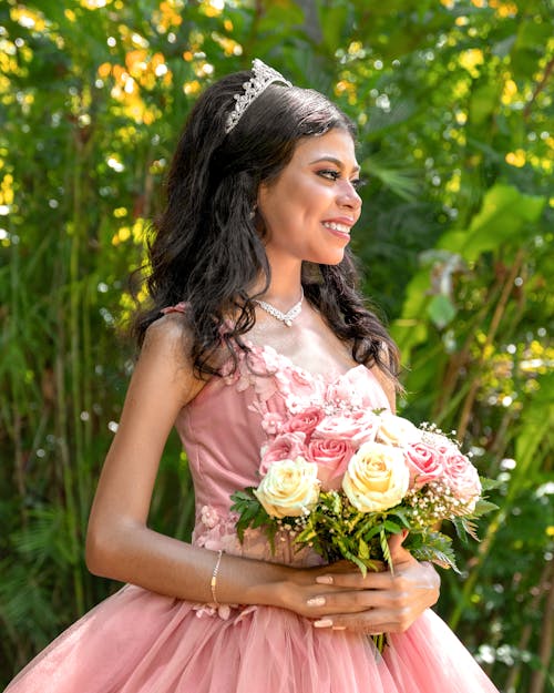 Young Woman in a Pink Princess Dress and a Tiara Standing Outside and Holding Flowers