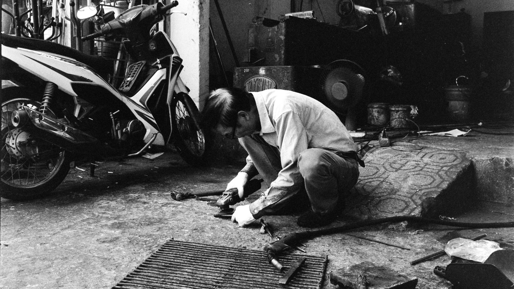 Black and white image of a mechanic working on a motor scooter in a workshop setting.