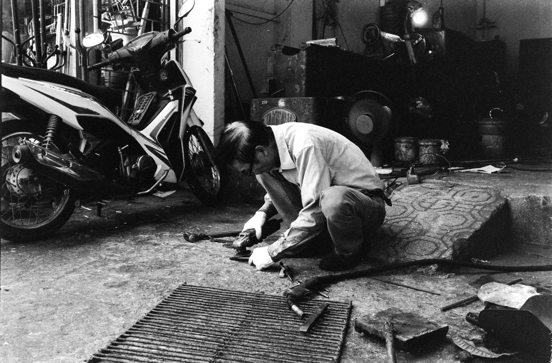 Black and white image of a mechanic working on a motor scooter in a workshop setting.