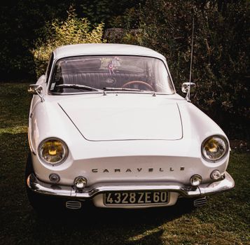 Front view of a vintage white Renault Caravelle parked outdoors with foliage background.