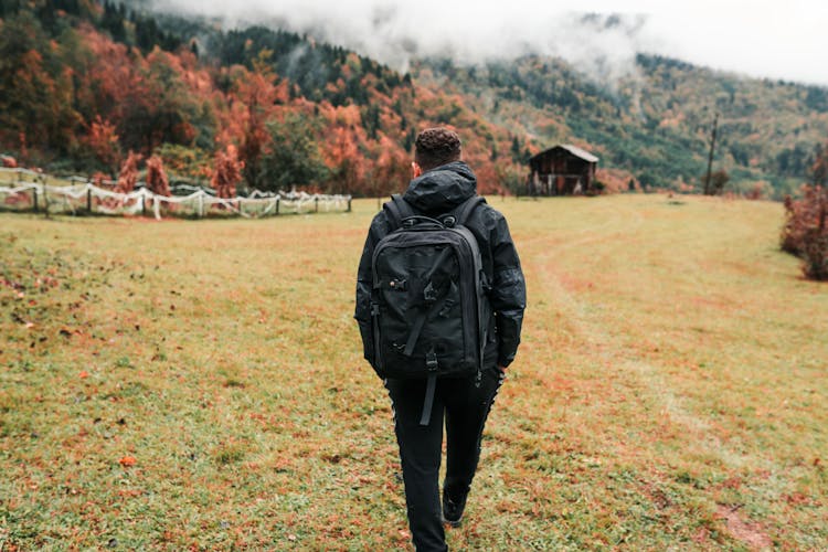 Back View Of A Man With A Backpack Walking On A Meadow In A Valley 