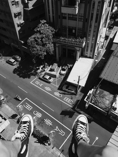 Grayscale Photography Man Wearing All Star Converse High Tops Below in Street and Building