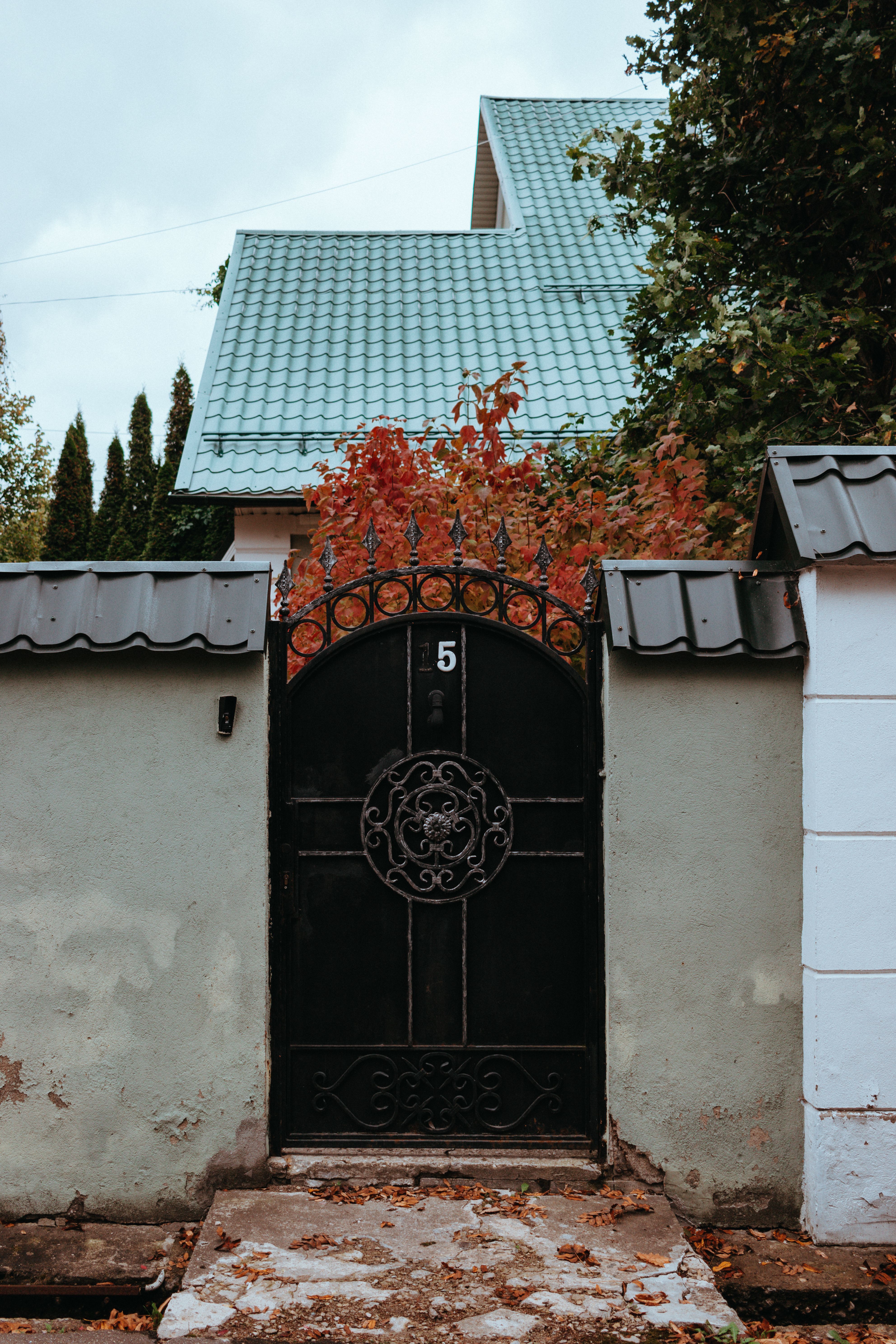 Charming entrance with black wrought iron gate and autumn foliage, leading to a house with green roof.