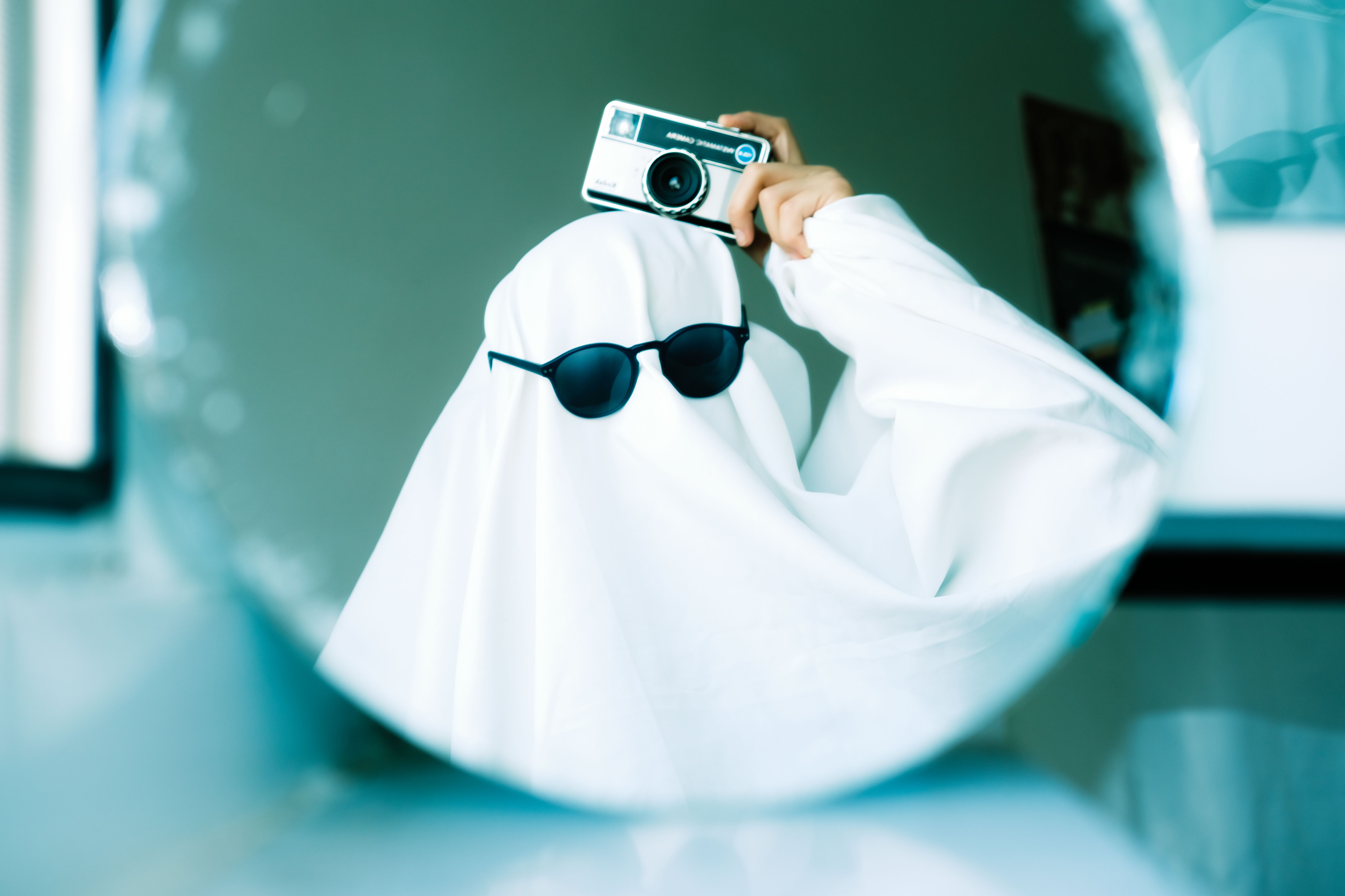 A person dressed as a ghost taking a selfie in the mirror with sunglasses on. Whimsical and fun.