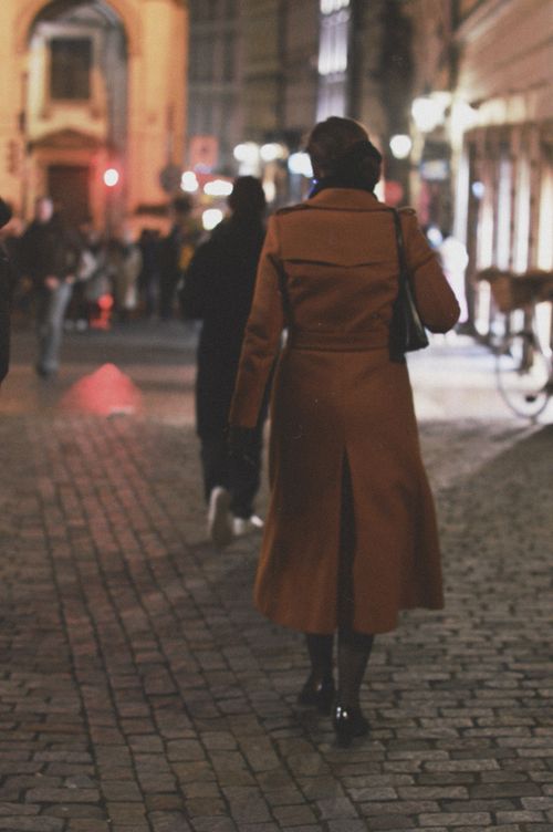 Free A woman in a brown coat walks on a cobblestone street at dusk in an urban setting. Stock Photo