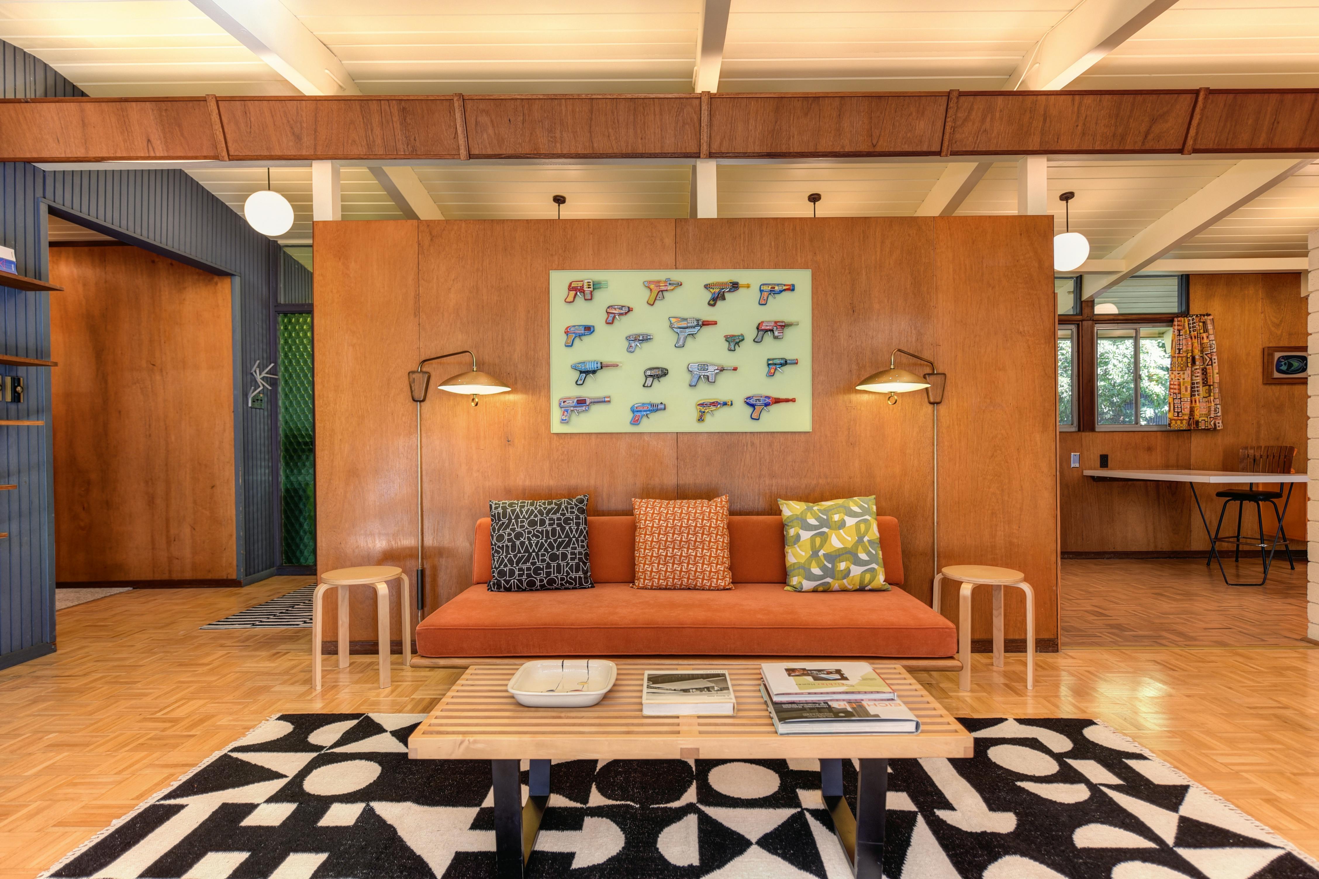 Mid-century modern living room with orange sofa, wood-paneled wall, and colorful ray gun pop art above couch