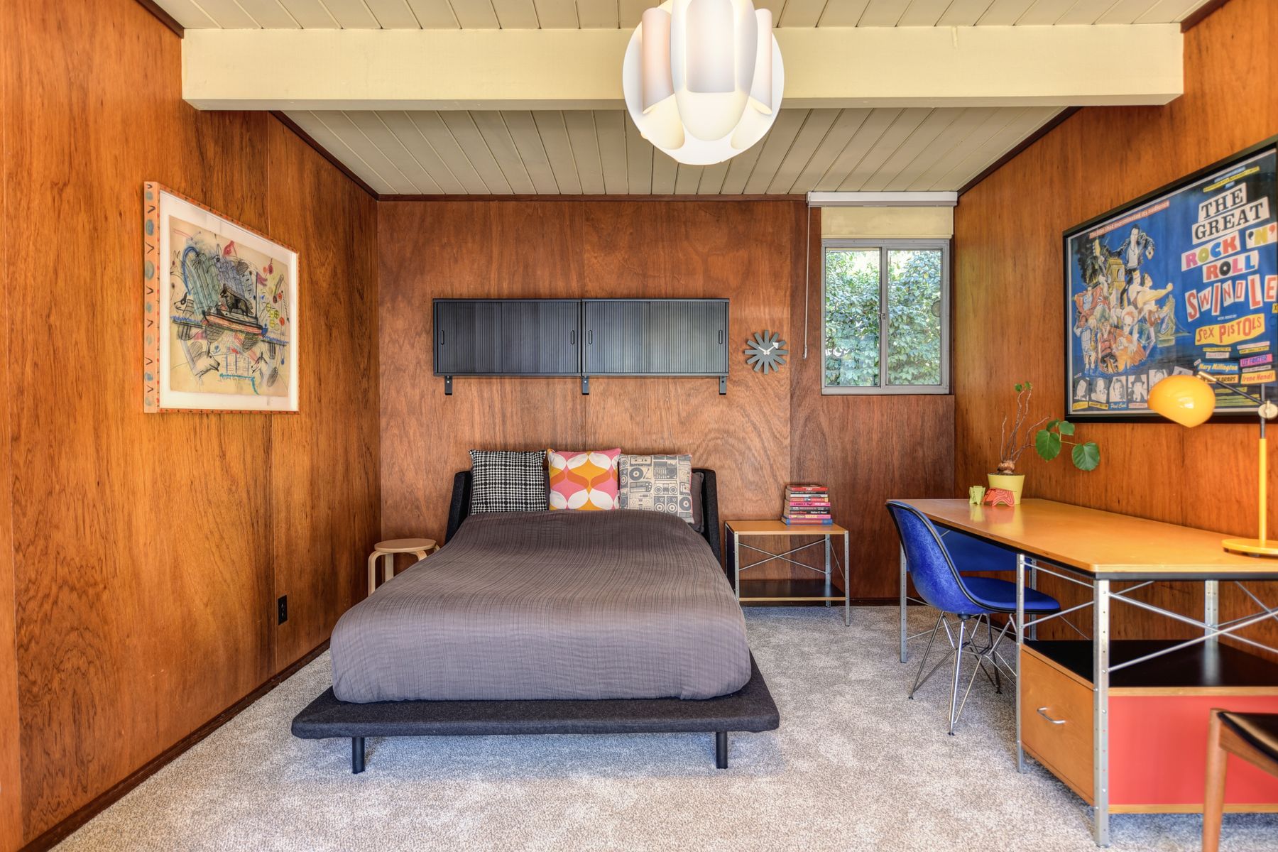 Warm mid-century modern bedroom with a low wood bed, wood-paneled walls, compact desk, rug and broad window