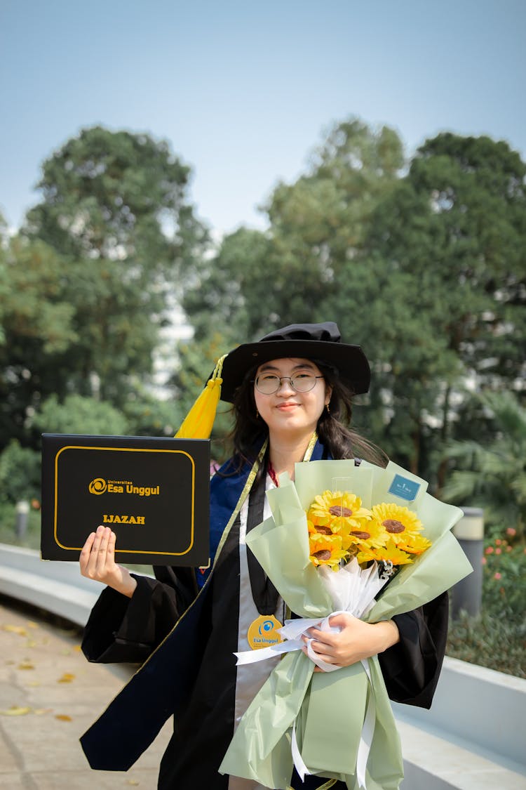 Young Woman In A Graduation Gown Holding Flowers And Diploma 