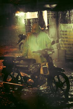 A man in a raincoat stands with his motorbike on a rainy night in the city.