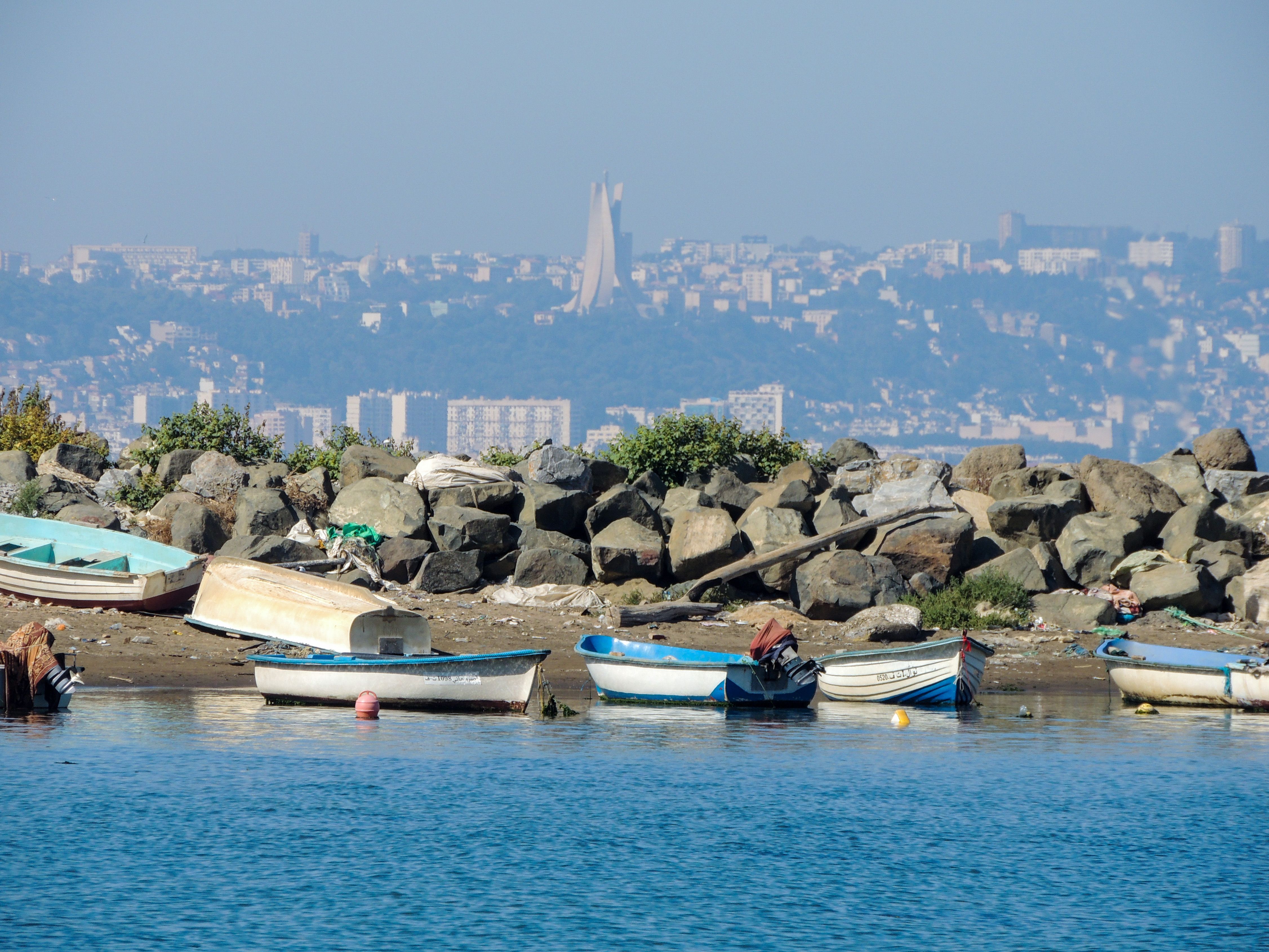 Boats moored along Bordj El Kiffan shore with Algiers cityscape in background, highlighting Maqam Echahid monument.