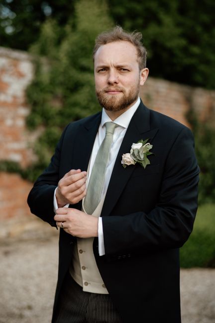 A refined man in a formal suit with greenery and a brick wall backdrop, exuding sophistication.