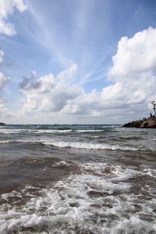 Free Tranquil coastal scene with waves crashing and a lighthouse in view under a blue sky. Stock Photo