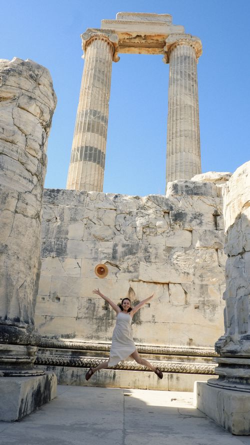 Free Young woman joyfully jumps in ancient temple ruins with towering stone columns under a clear blue sky. Stock Photo