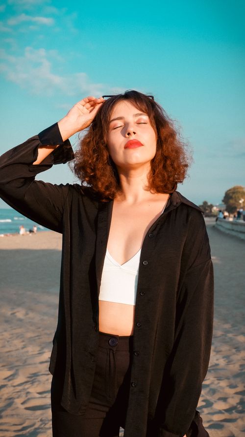 Free A woman with curly hair enjoys the sun on a sandy beach, wearing a black shirt and white top. Stock Photo