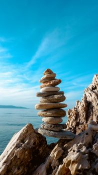 A stack of balanced stones on a rocky seashore with a clear blue sky and sea in the background.