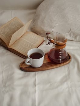 A warm cup of coffee on a bed with an open book and a coffee carafe, perfect for relaxation.