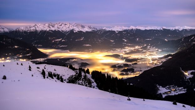 A stunning winter view of the Dolomites with snow-covered peaks and glowing valley lights at twilight.