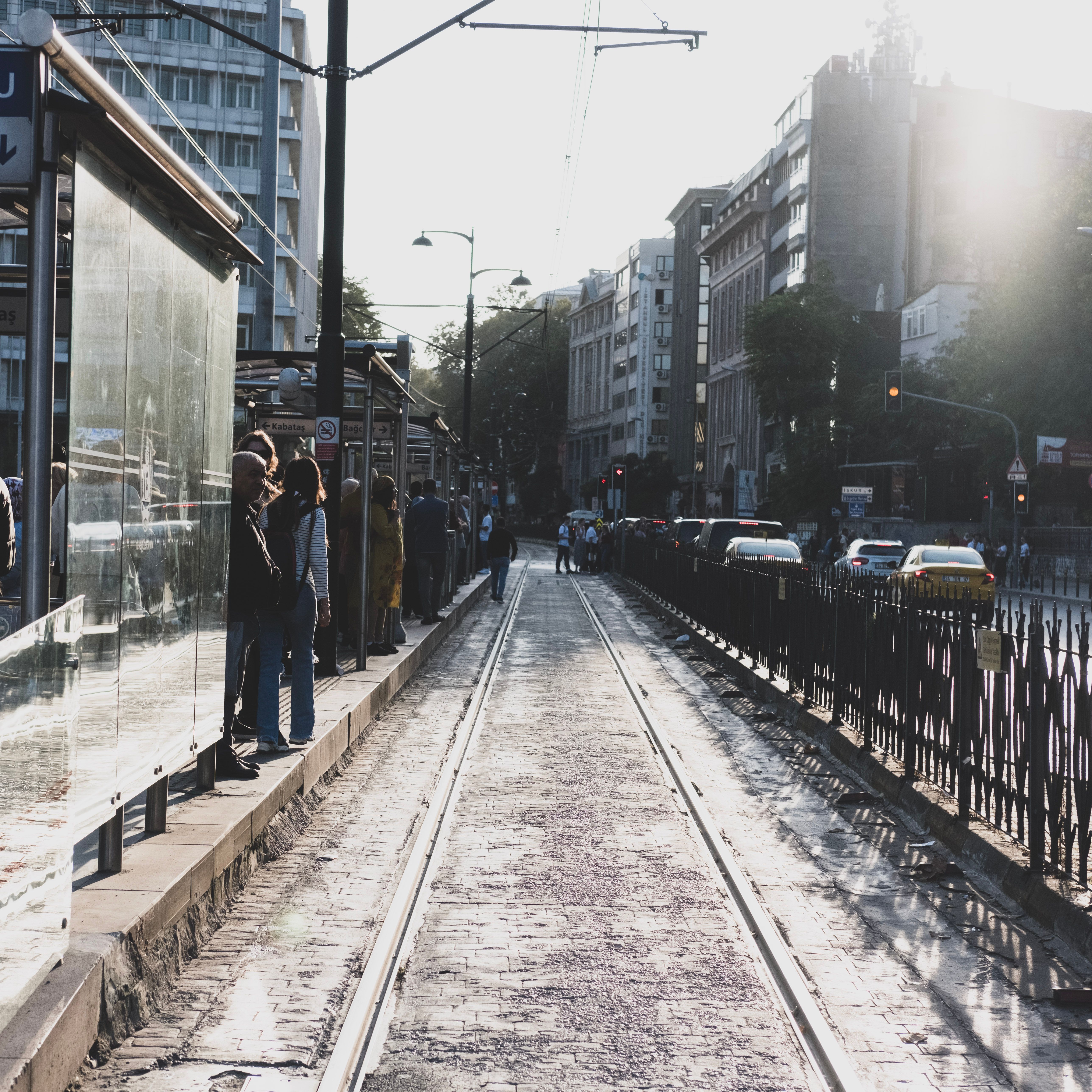 People waiting at a busy city tram station during sunset with urban architecture in the background.