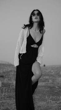 A stylish woman poses confidently in a black dress and sunglasses in a desert setting.