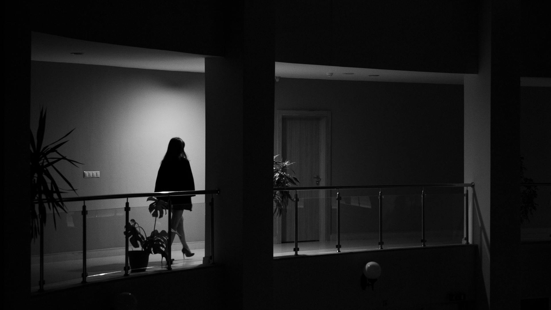 Silhouetted woman walking along a dimly lit balcony corridor at night.