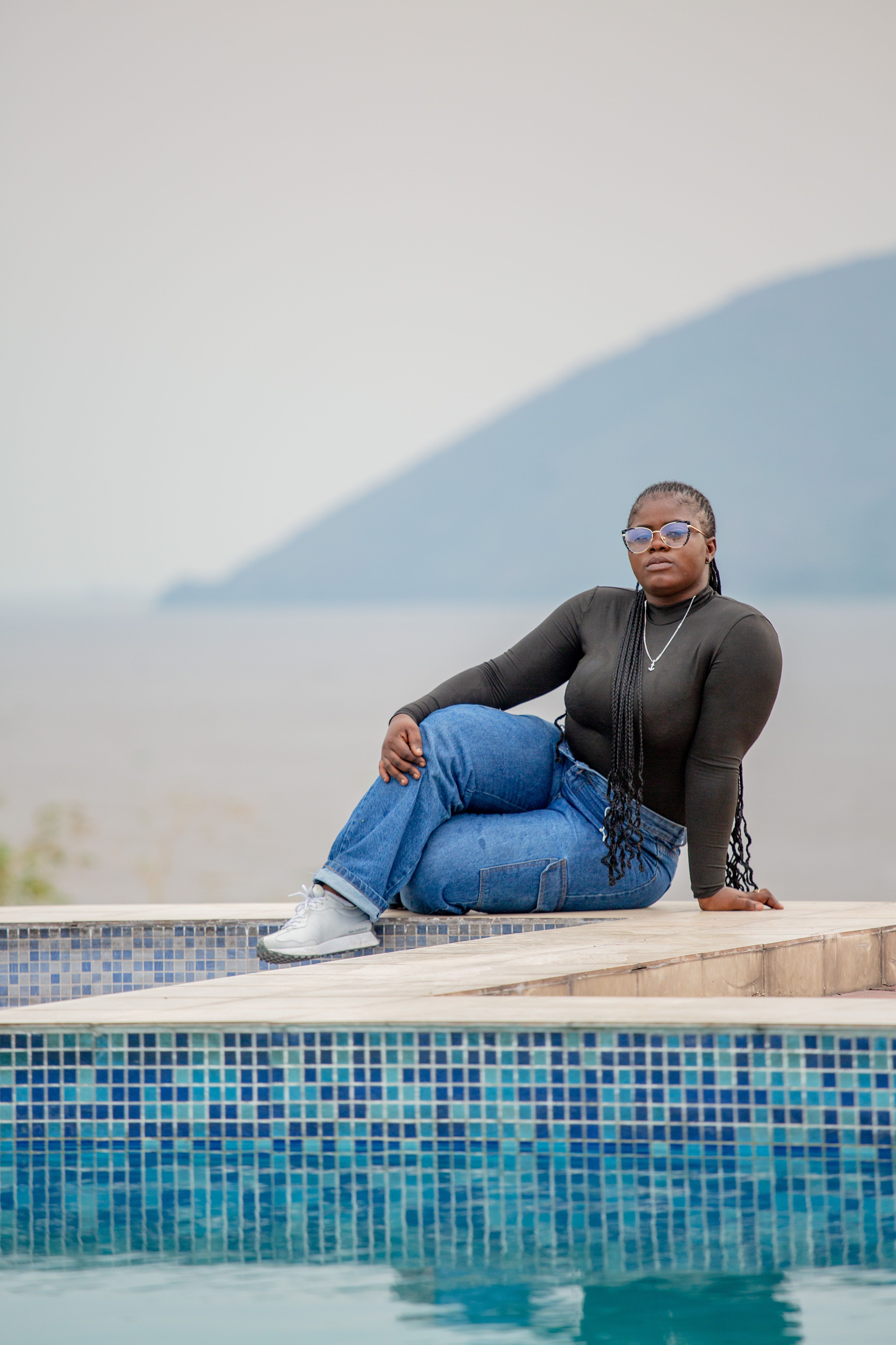 An elegant woman in sunglasses posing by a pool in Kinshasa, showcasing a relaxed summer fashion vibe.