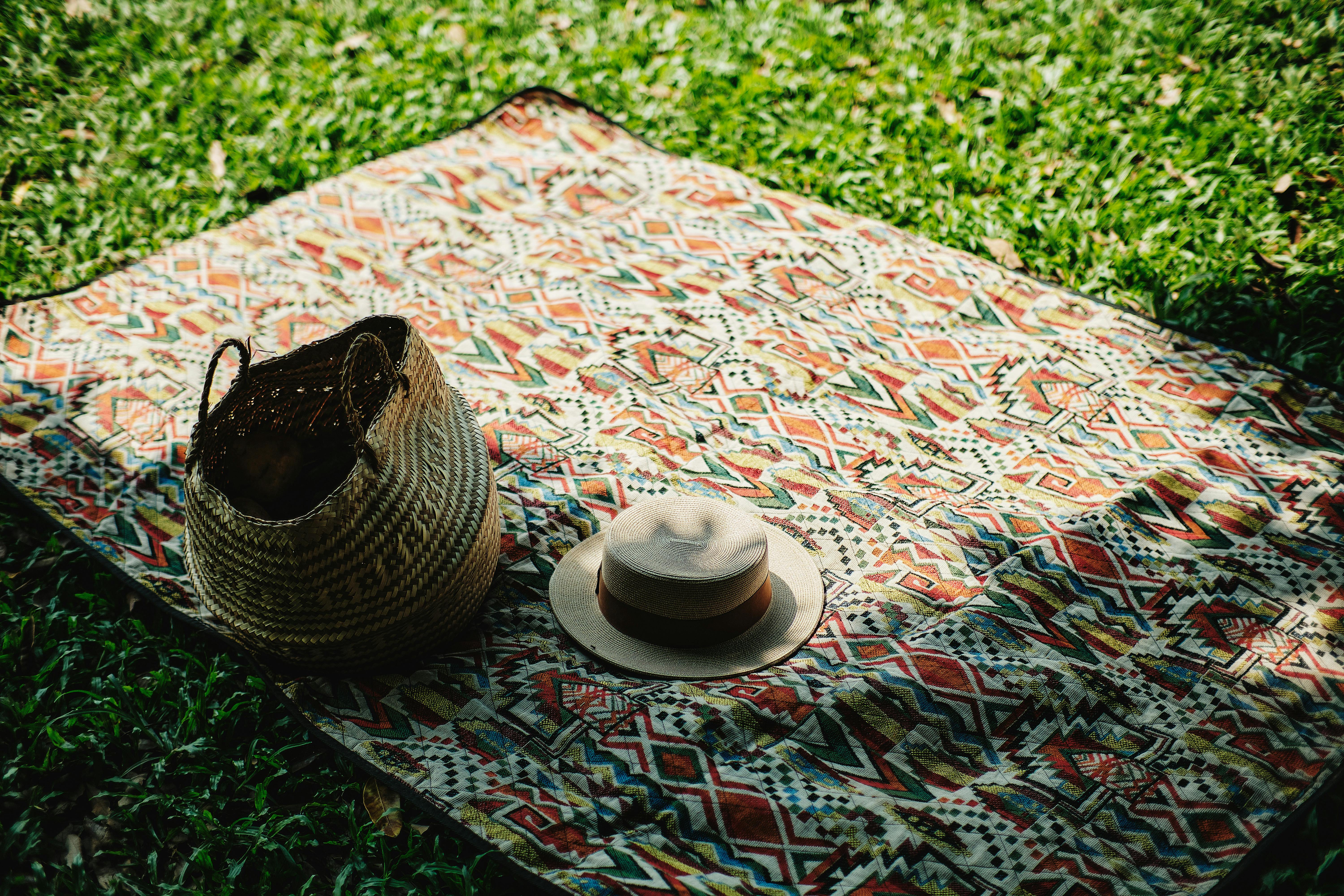 Colorful picnic setup with a hat and basket on grass under sunlight.