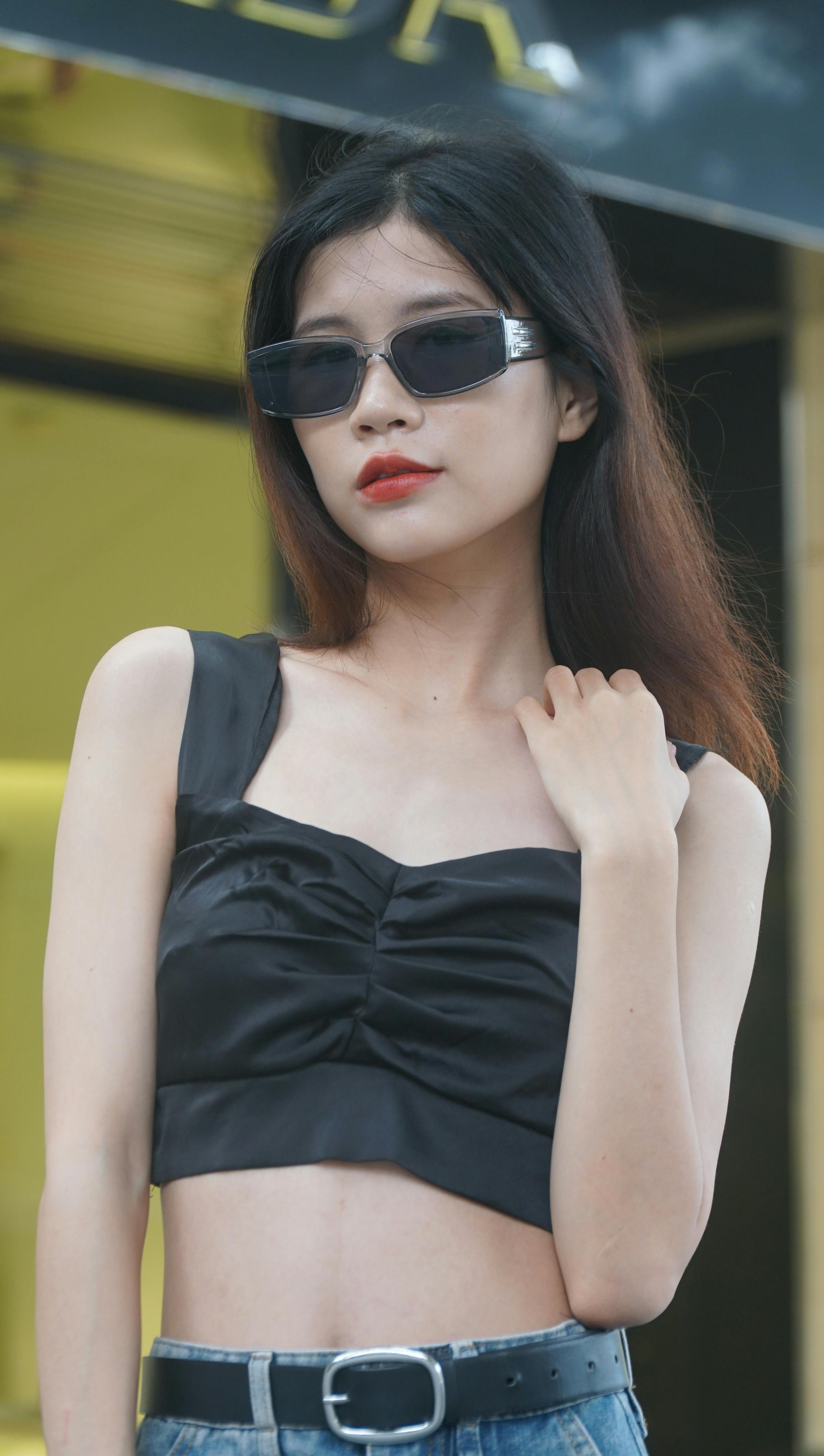 Fashionable woman with black hair wearing sunglasses and a black top, posing confidently outdoors.